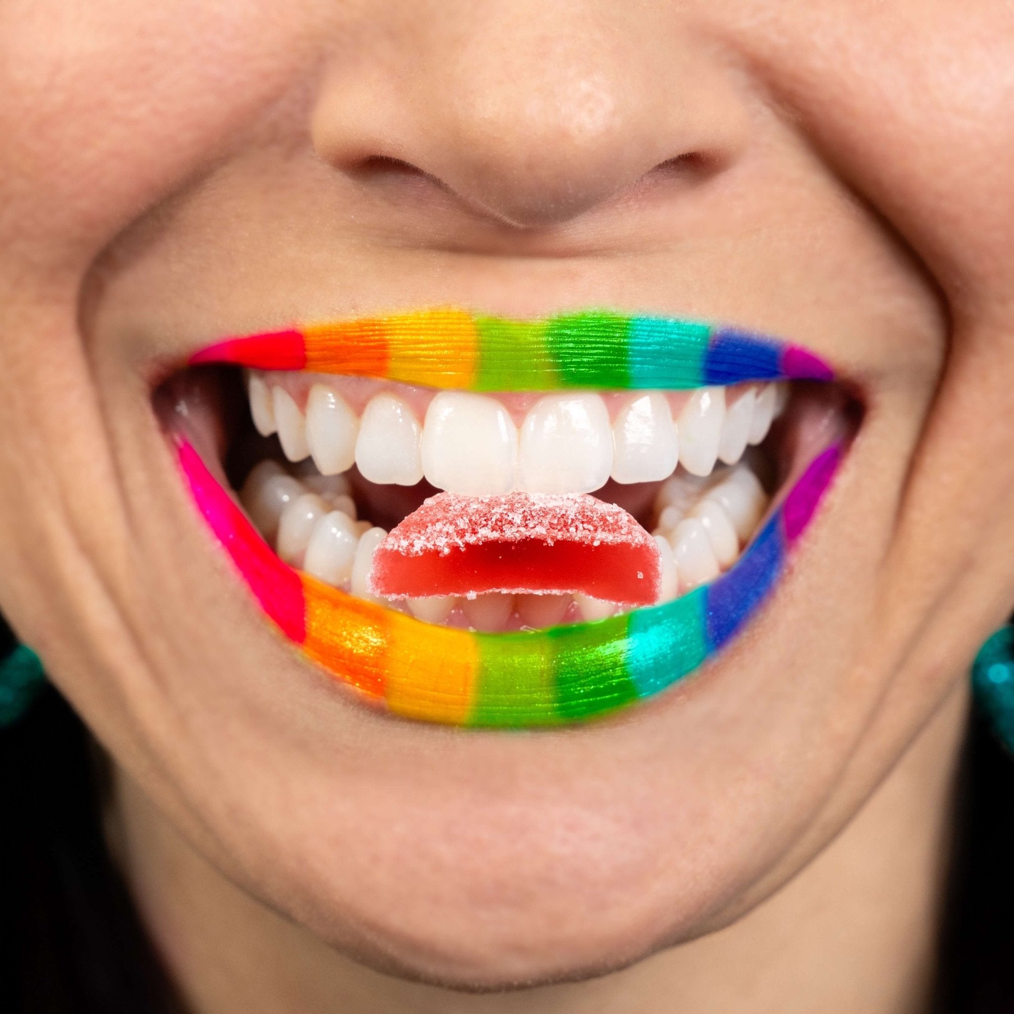 Nothing But Hemp Zkittles hemp-derived THC indica gummies with rainbow lip styling inspired by the package holding a split gummy between the teeth against a pink gray background in a lifestyle scene