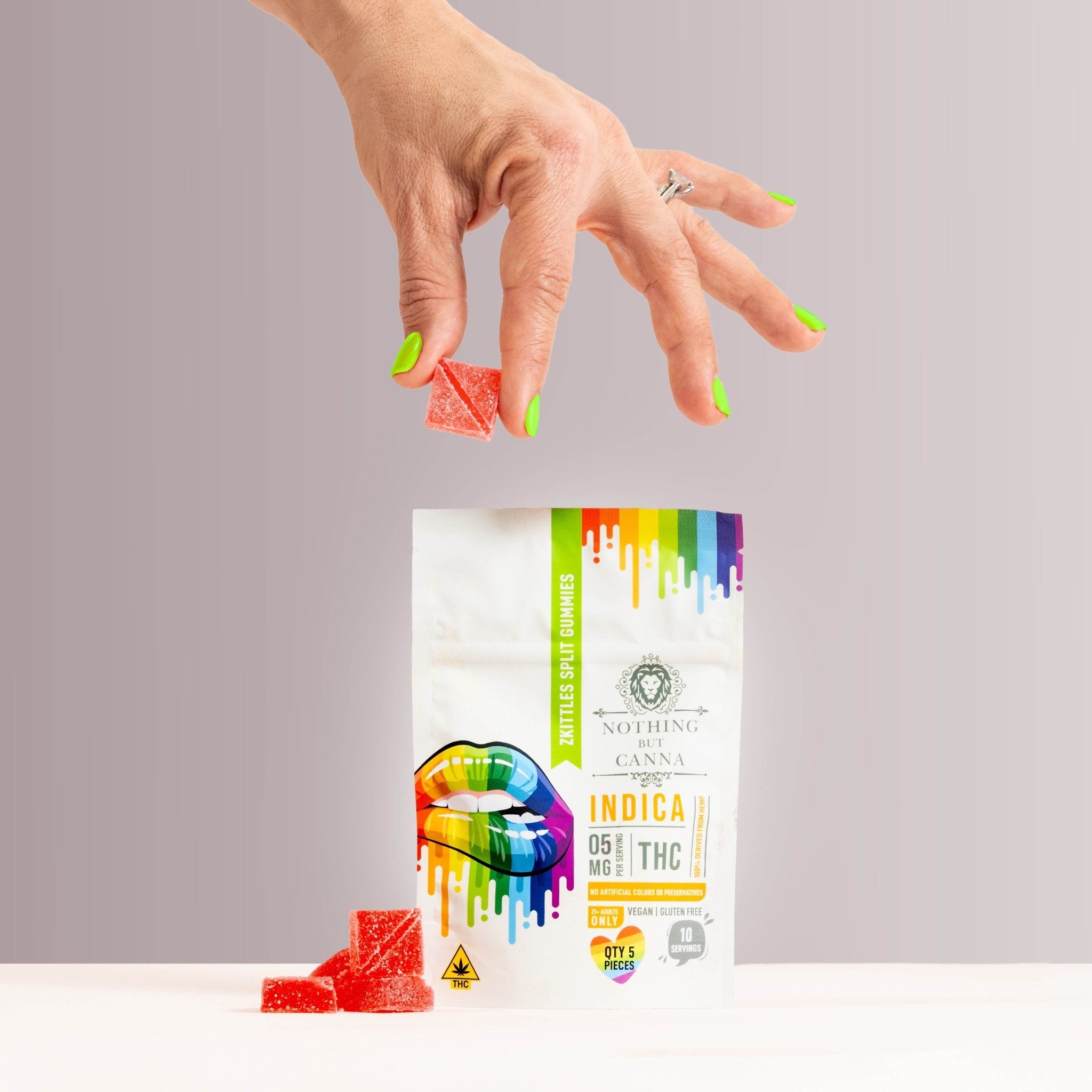 Nothing But Hemp Zkittles hemp-derived THC indica gummies package on a pink gray surface with a hand with green fingernails holding a gummy above the bag and a handful of gummies placed beside the product in a lifestyle scene