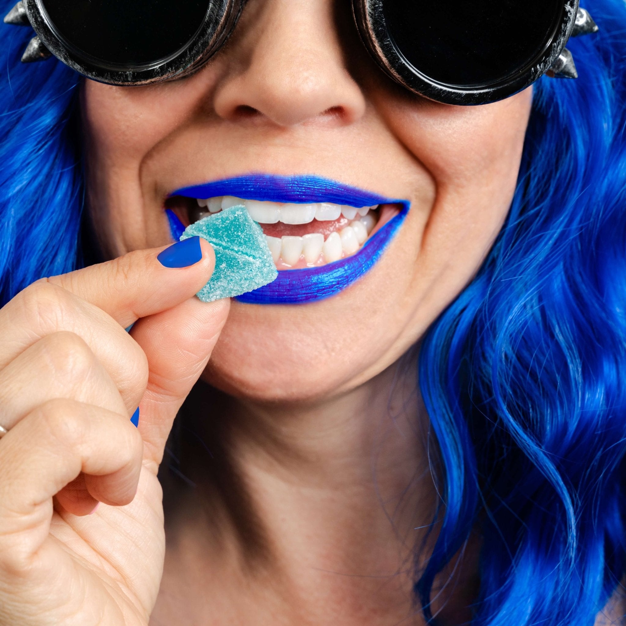 Person styled to match Blue Dream packaging with steampunk goggles, blue wig, blue lipstick, and blue fingernails about to place a hemp-derived delta-9 THC sativa gummy from Nothing But Hemp into their mouth while holding the product bag featuring the illustrated character