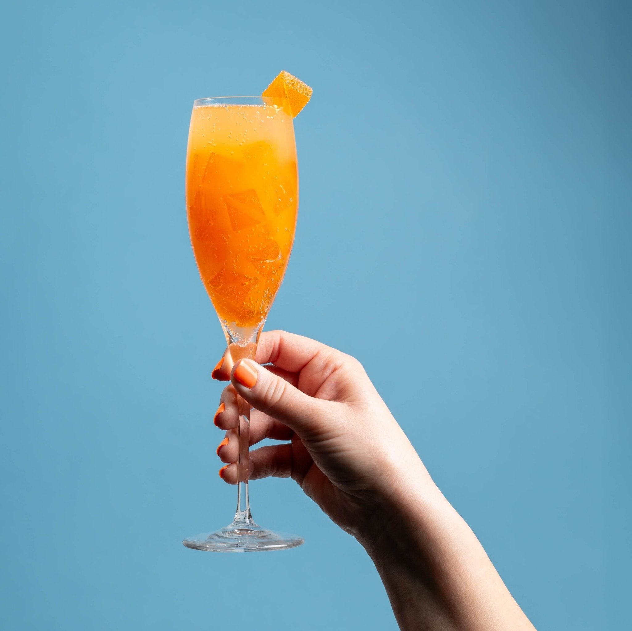 Nothing But Hemp Mimosa hemp-derived THC and CBD sativa gummies shown in a champagne glass filled with orange gummies and champagne, with a gummy garnished on the rim, held by a hand against a sky blue background in a lifestyle scene