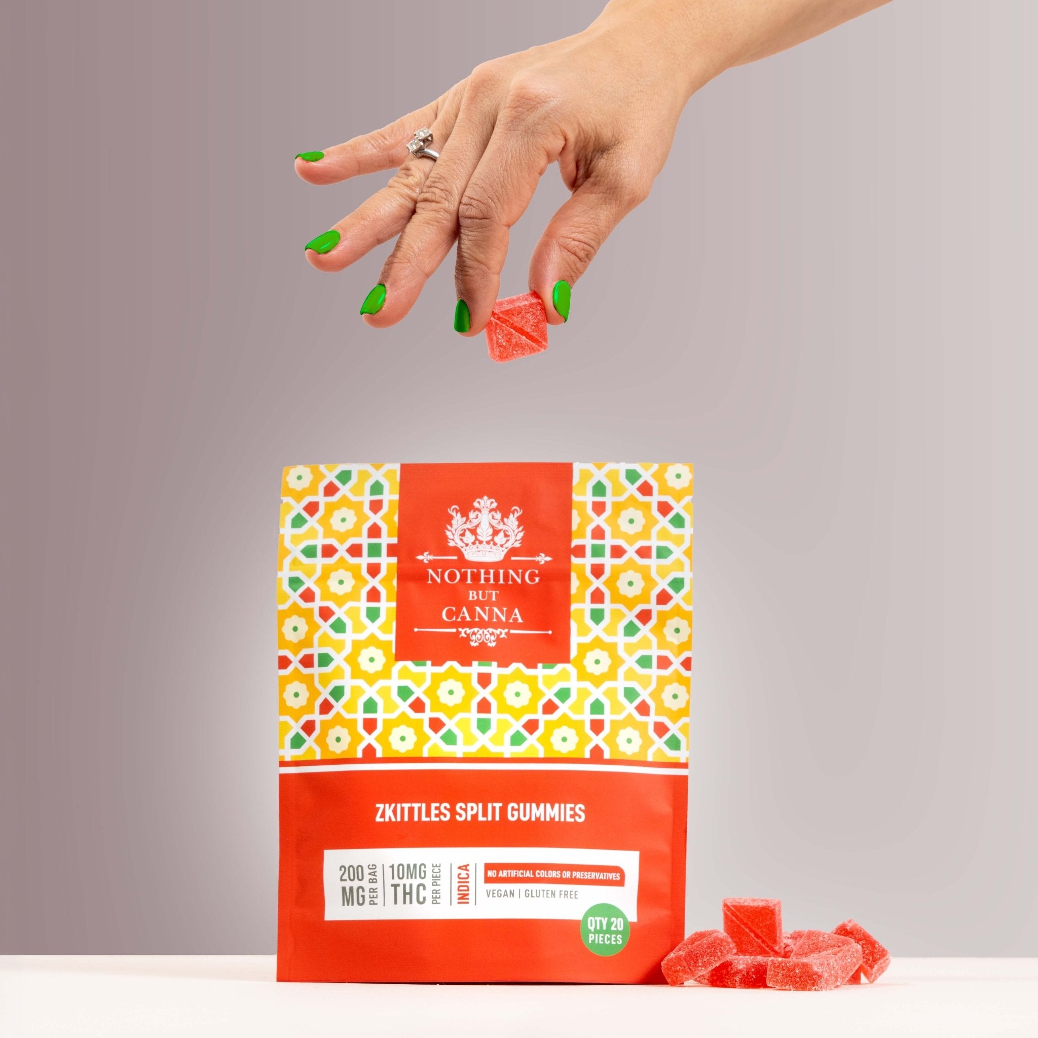 Nothing But Hemp Zkittles hemp-derived THC indica gummies package on a pink gray surface with a hand with green fingernails holding a gummy above the bag and a handful of gummies placed beside the product in a lifestyle scene